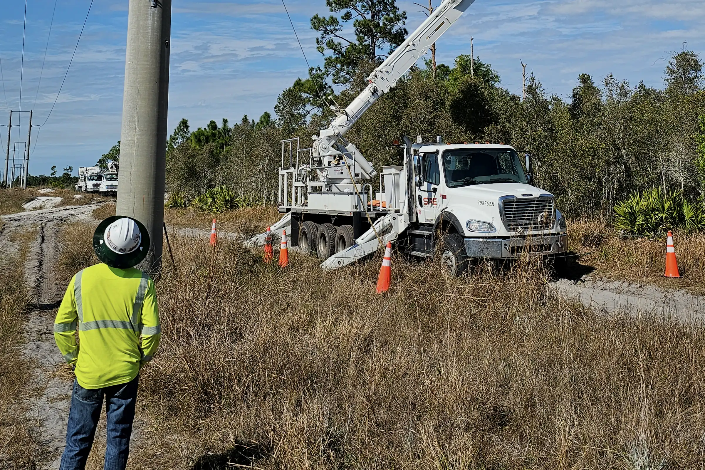 Ground worker in a hard hat and hi-vis shirt watching an SPE bucket truck lift at a transmission pole